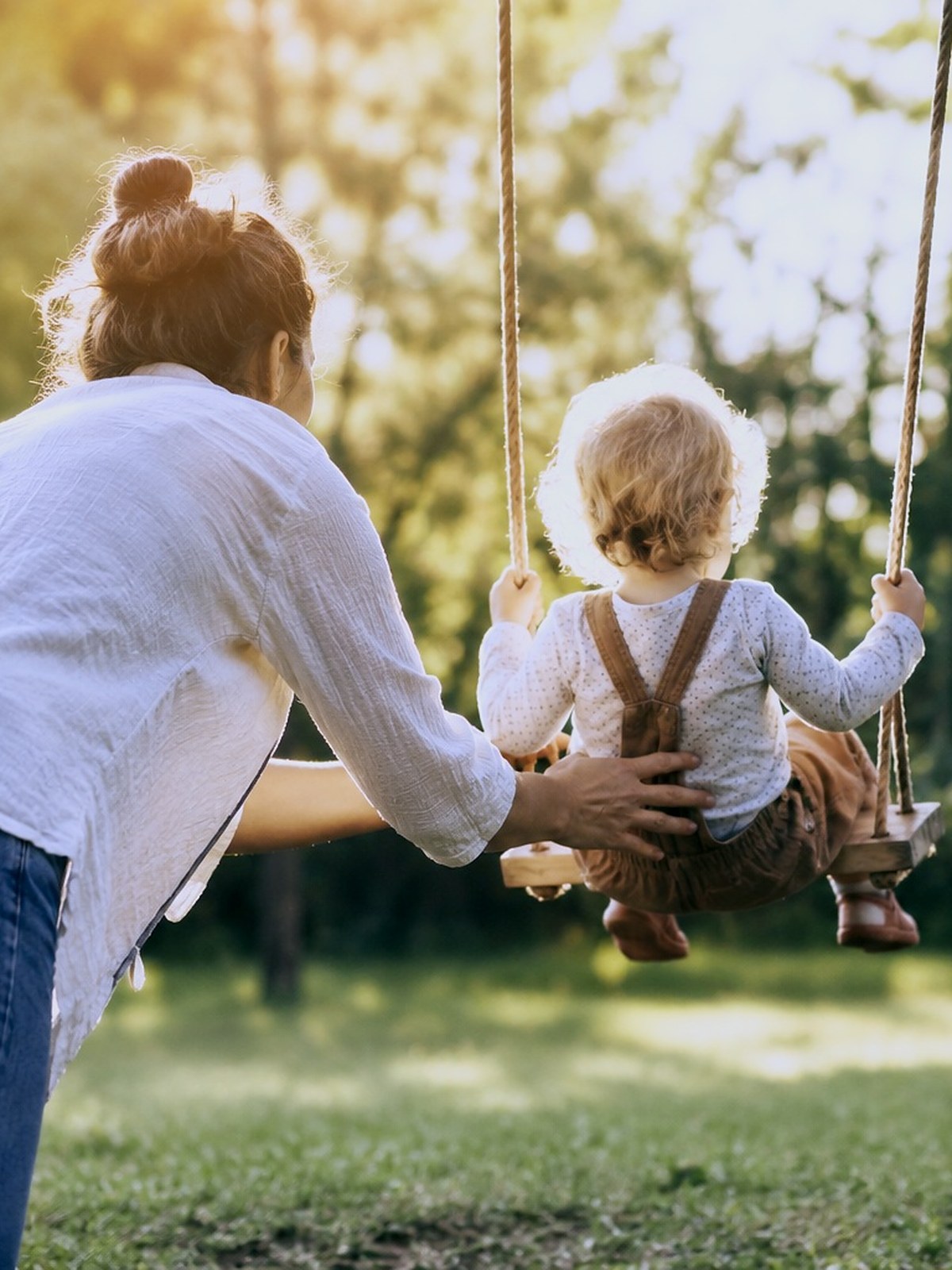 Eine Mutter schubst ihr zweijähriges Kind auf einer Holzschaukel an, beide von hinten gesehen.