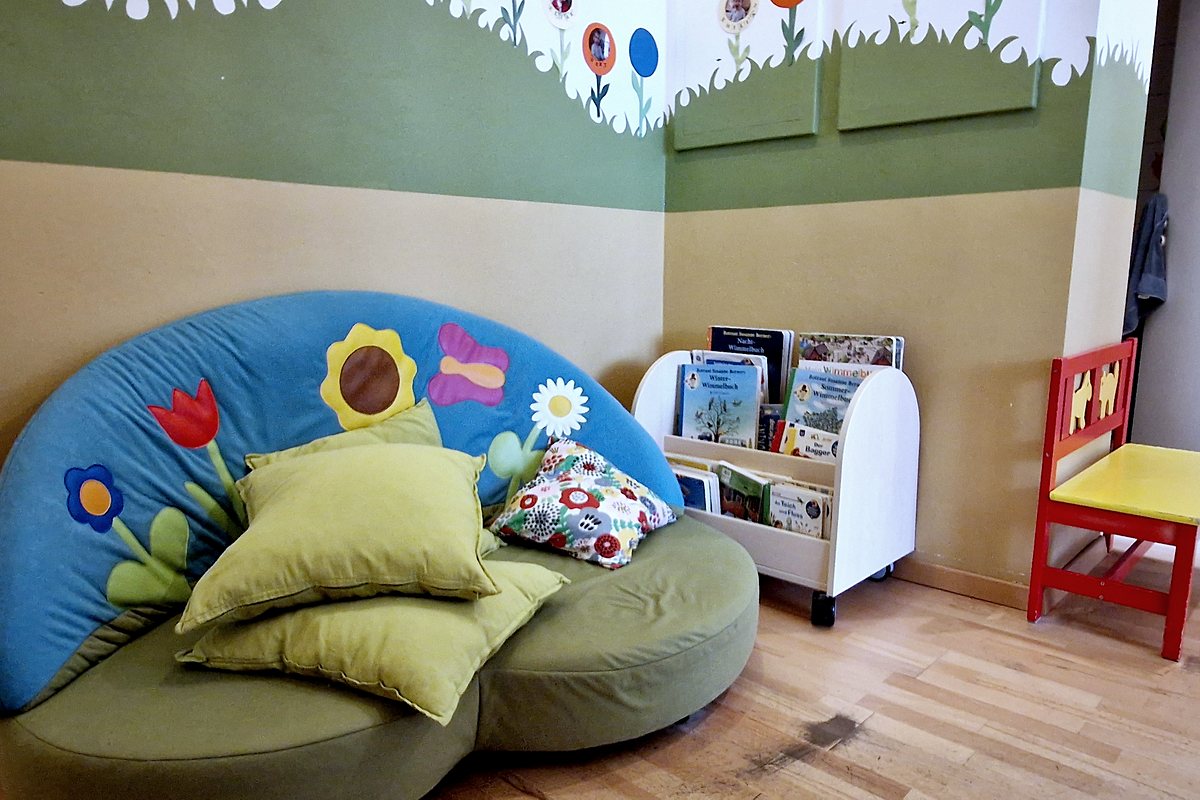 Gemütliche Leseecke mit Kindercouch, Kissen und einem Bücherwagen voller Kinderbücher.